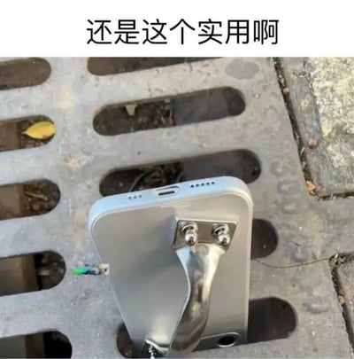图片展示了一部银色智能手机被一个金属支架固定在带有长条形孔洞的井盖表面，支架通过两颗螺丝与手机背面连接，手机底部有一根绿色接头的电缆延伸出来。图片顶部配有文字“还是这个实用啊”，整体场景呈现出一种民间创意的实用装置，可能用于手机的固定放置或外接设备使用，透露出对这种简易但有效的解决方案的赞赏。