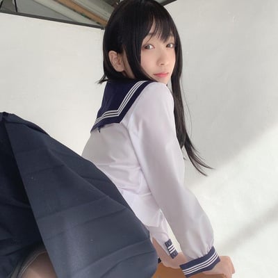 图中是一位身着经典款JK水手服的长发女生，她背对着镜头侧身回头，眼神柔和地看向镜头，姿态温婉。上衣为纯白色，搭配带有三道白色条纹的藏青色水手领，下身是同色系藏青色百褶裙，背景是简洁干净的白色墙面，整体画面充满清新治愈的校园氛围，风格甜美温柔。