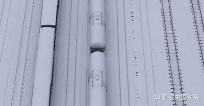 这是一张高空俯拍的冬日雪景实拍图，画面主体是被皑皑白雪完全覆盖的高铁接触网配套设备，设备的金属箱体上的固定装置与积雪搭配，恰好形成了类似笑脸的图案。两个上下排列的箱体，在视角和图案的加持下，看起来如同两个面对面的带笑脸的“小人”，充满趣味。画面右侧是同样被积雪覆盖的铁路轨道相关设施，原本冰冷硬核的铁路运维设备，在大雪的装扮下显得格外可爱，为冬日的铁路场景增添了一份温馨趣味。