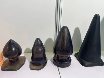 图片展示了四个黑色锥形物体并排摆放在白色平面上。这些物体呈现出不同的形状和大小，从左到右依次为：一个底部较宽、顶部圆润的锥形物体；一个形状较为规整的锥形物体；一个底部较宽、顶部略尖的较大锥形物体；以及一个高度最高、形状最尖锐的锥形物体。所有物体表面光滑，具有一定的反光效果，背景为白色墙壁，整体画面简洁干净。