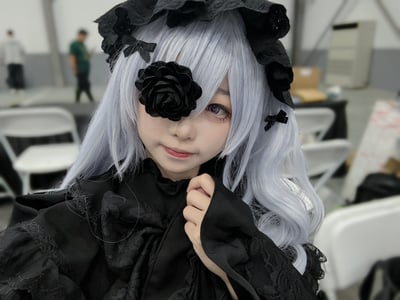 这是一张真人cosplay实拍照片，出镜者还原了哥特风格的二次元角色造型：佩戴银灰色微卷长假发，头上装饰黑色蕾丝荷叶边发箍，发丝上别有数个黑色小蝴蝶结，左侧眼部以黑色布艺玫瑰作为遮挡，身穿带有蕾丝花边设计的黑色哥特风裙装。出镜者对着镜头面露柔和微笑，右手抬起轻抓自己的衣领，背景是开阔的室内场馆，可见白色折叠椅和模糊的路过人员，符合漫展活动的场地特征。