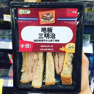 图片中一只手拿着一盒超市售卖的三明治包装，主体为黑色托盘+红色标签设计。顶部有绿色“ohyama plant”标志和三明治插图，正面醒目大字“地板三明治”搭配副标题“做的时候不小心掉了哈哈”，左下角红色“半价！”标签格外显眼，下方标注“烟熏鸡胸和火腿(很抱歉 原谅啦)”，右下角有眯眼微笑的黄色表情符号。侧面印有英文储存提示“KEEP REFRIGERATED OR FROZEN”及净含量“6.3 OZ (179g)”。通过“地板”这一荒诞命名和“掉地上半价”的幽默文案制造笑点，模拟超市奇葩商品场景。