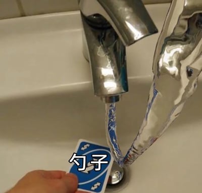这是一张利用视觉错位打造的趣味静态图，场景设置在洗手池前：水龙头流出一段水流，旁边用锡纸制作出和水流视觉衔接的造型，一只手拿着一张带有+4标识的类似UNO的卡牌放在水流下方，图片上标注了「勺子」字样。通过视角的巧妙搭配，让水流、锡纸和卡牌组合出仿佛「勺子」在承接水龙头水流的错觉，是充满创意的趣味视觉小创作，用来制造搞笑、新奇的视觉效果。