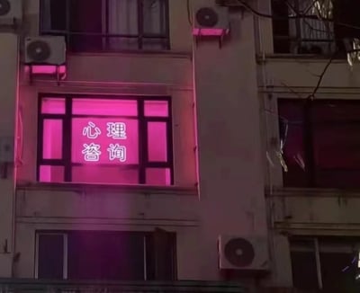夜晚的居民楼外墙场景，一栋老式建筑的某扇窗户被明亮的粉色灯光完全照亮，窗玻璃上清晰显示着白色的“心理咨询”四个汉字。建筑外墙上安装有空调外机，部分树枝从画面右侧伸入，整体呈现出一种静谧又略带神秘的夜间氛围。