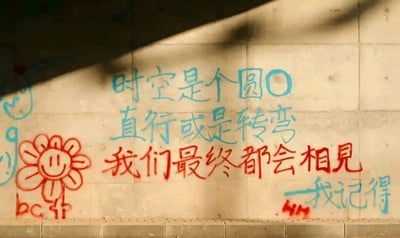 这是一张拍摄城市墙面涂鸦的静态照片，墙面为水泥材质，左上角有部分阴影遮挡。墙面上的涂鸦使用蓝色和红色喷漆创作，包含文字与手绘图案：蓝色喷漆书写“时空是个圆○ 直行或是转弯 我记得”，红色喷漆书写“我们最终都会相见”；左下角有红色手绘的笑脸太阳花图案，花的下方配有红色的“bc jf”字样，右下角还有红色的“MM”标记。整体涂鸦风格质朴治愈，通过“时空是圆”的比喻，传递出无论人生选择何种轨迹，最终都会与珍视之人重逢的温暖期许，是充满人情味的民间街头创作。