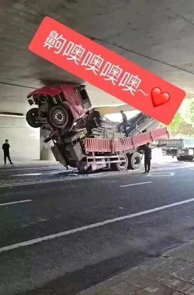 一张交通事故现场的照片被编辑成搞笑梗图。图中一辆红色卡车疑似因超高撞击立交桥底部，车头向上翘起，车身倾斜。照片上方添加了一个红色标语牌，上面写着“齁噢噢噢噢~❤️”，用夸张的语气词表达惊讶或感叹，与事故现场形成幽默对比。桥下有两名身穿深色衣服的人，一人站在卡车旁，另一人似乎在观察情况。