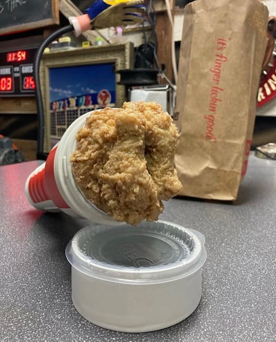 一张展示超大炸鸡块的趣味照片：一块金黄色的炸鸡从红白条纹的杯子里满满地溢出来，几乎要掉落到下方的透明塑料盖子上。背景中可见印有经典广告语'it's finger lickin' good'的棕色纸袋（疑似肯德基包装），周围环境像是快餐店后厨或家庭厨房，有显示11:54的数字时钟、带边框的相框和各类厨房用具，整体画面通过夸张的炸鸡尺寸营造出惊喜感和视觉冲击。