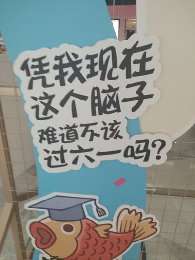 这是一张商场或公共场所摆放的彩色卡通宣传牌，牌子上有大字标语：“凭我现在这个脑子，难道不该过六一吗？”配图是一只戴着学士帽的卡通鲤鱼，表情呆萌搞笑。整体画风轻松幽默，传递了成年人因压力过大、脑袋“变幼稚”的自嘲心态，表达大家为生活和学业所累，调侃自己已经没有“成年人的脑子”，很适合用来调侃职场、学生党等在压力下渴望“返老还童”的心情。

**梗点解析**：
- “过六一”指儿童节，通常只属于小朋友，被成年人“抢过”，是当下网络热门梗。
- “凭我现在这个脑子”自嘲思考能力退化，带有搞笑和自黑意味。
- 图中的鱼通常指“咸鱼”，有“颓废、不想奋斗”的网络含义。