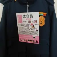 这是一张竖版实拍照片，主体为一件深蓝色外套，外套上悬挂着一张带有粉色边框的「试穿品」提示牌。提示牌采用图文结合的形式，标注了试穿样衣的多项注意事项，包括试穿前确认款式尺码、避免化妆喷香水弄脏衣物、试穿后归位、污渍需按原价购买、爱护商品避免损坏等内容，场景应为线下服装店，用于规范顾客的试穿行为。