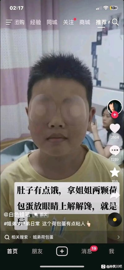 这是一张竖屏的社交媒体截图，画面主体是一位短发小男孩，他的双眼位置贴着两个圆形眼贴，被配文调侃成“荷包蛋”；背景里能看到一位女性（姐姐）正专注看手机。配文写道“肚子有点饿，拿姐姐两颗荷包蛋放眼睛上解解馋，就是玩”，还带有#姐弟 #姐弟日常 这个荷包蛋有点粘人 的话题标签，以玩梗的方式将眼贴比作荷包蛋，打造出姐弟间的搞笑日常内容，充满生活化的趣味。截图保留了抖音平台的界面元素，包括顶部的导航栏、右侧的点赞、评论、关注按钮，底部的首页、朋友等导航栏，右下角还有百度贴吧孙笑川吧的水印，说明该内容被搬运到贴吧进行分享。