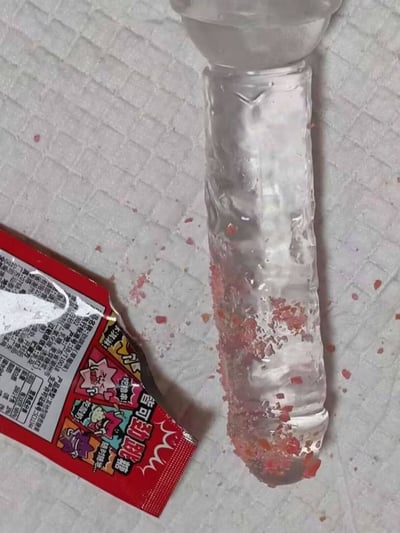 这张实拍图的背景是带有格纹的白色桌面，左侧放着一包已经拆开的红色包装劲跳糖，右侧摆放着一根透明的冰棍，冰棍的下半部分沾满了红色的跳跳糖颗粒，桌面上也散落了少量跳跳糖碎粒，展示了将跳跳糖搭配冰棒食用的新奇趣味吃法。