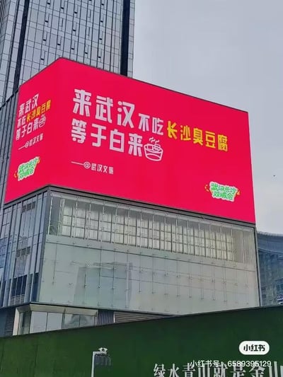 这是一张户外实拍图，画面主体是一块安装在现代玻璃幕墙建筑外立面上的大型红色电子广告牌。广告牌以鲜艳红色为背景，用醒目的白色和黄色字体呈现宣传语“来武汉不吃长沙臭豆腐等于白来”，下方标注“——@武汉文旅”，同时配有“武汉长沙双城会”的绿色卡通标识。背景是高层现代写字楼，广告牌下方是绿色施工围挡，围挡上印有“绿水青山就是金山”的字样，画面右下角带有小红书平台的水印及发布者的账号信息。这是武汉与长沙的文旅联动宣传内容，打破常规本地宣传思路，以反向推荐邻城特色美食的幽默方式制造话题，吸引游客关注双城旅游。