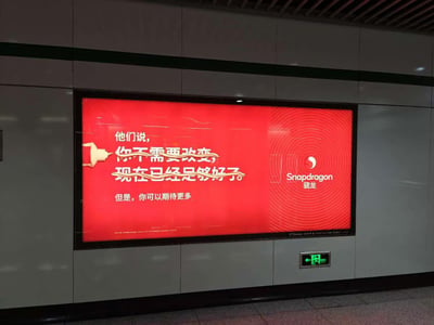 这是一张拍摄于地铁站内的照片，画面主体是一块安装在灰色墙面的红色背景骁龙品牌广告牌。广告牌原本完整文案为：“他们说，你不需要改变，现在已经足够好了。但是，你可以期待更多”，巧合的是广告牌左侧墙面伸出的浅色装饰结构，形成两条横带刚好划掉了“你不需要改变”和“现在已经足够好了”两句文案，仅保留“但是，你可以期待更多”，形成了巧妙的视觉错位效果，传递出鼓励突破进取、追求更好的含义，广告牌右侧印有骁龙（Snapdragon）的品牌logo，广告牌下方可见一块绿色的地铁站安全出口指示牌。