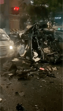 这是一段记录深夜城市路口严重交通事故的动态图，画面中多辆汽车在事故中遭受重创：一辆轿车的车头与车身侧面完全变形，车身部件破碎散落，路面上铺满了各类车辆残骸与碎片；另有一辆小型货车车头也发生严重变形，整体损毁状态触目惊心。现场可见亮起的交通信号灯，环境为夜晚的城市道路，直观展现出这场车祸的强大冲击力，场景十分惨烈。