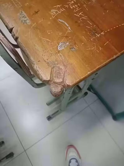 这是一张在教室拍摄的实拍照片，画面中的旧木质课桌因长期使用，桌角的木头出现磨损变形，搭配桌面的划痕痕迹，从当前拍摄角度看，桌角与桌面痕迹组合形成了类似大蜘蛛倒挂在桌沿的视觉错位效果，极具趣味性。桌面还留有学生刻写的“到此一游”“大可”等字迹，课桌的金属支架、教室的瓷砖地面也出现在画面中，整体充满校园日常的生活感。