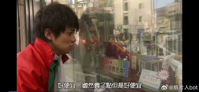 图片中一名身穿红色夹克的男子正站在玻璃橱窗前，专注凝视着橱窗内陈列的玩具或模型商品。橱窗玻璃反射出街道背景与建筑物，画面下方配有中文字幕：“好便宜 雖然貴了點但是好便宜”，形成鲜明的矛盾心理表达——嘴上说着贵，却仍觉得划算。男子表情透露出对商品的渴望与纠结，整体场景充满生活化的幽默。