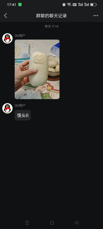 这是一张QQ群聊记录的截图，显示一位QQ用户在昨天17:14分享了一张照片。照片中一只手正拿着一个表面印有类似表情包脸图案的馒头，馒头造型方正，面部图案简洁，类似常见的搞怪表情包。背景中可以看到其他几个普通形状的馒头放在碗里，桌面铺着带有卡通图案的桌布。图片下方紧跟着用户发送的文字“馒头B”，整体呈现出群聊中分享生活趣事的轻松场景。