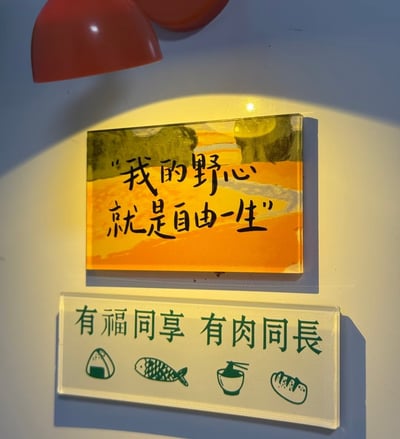这张图片拍摄了一面墙上的两块创意标志牌。上面的橙黄色牌子用手写体写着“我的野心就是自由一生”，表达追求自由无拘的人生态度，语气洒脱带有些许文艺气息。下方的白色牌子写有“有福同享 有肉同长”，并配有饭团、鱼、饭碗和面包的简笔画图案，巧用谐音梗（有“福”（福气）同享，有“肉”（快乐/实际好处）同长），搞笑中带点温馨，常用于朋友或团队共进美食的情境。整体风格温暖、明亮，适合餐厅、咖啡店或文艺空间装饰。

**谐音梗说明**：第二句“有肉同长”与成语“有福同享，有难同当”谐音反转，将“难”换成“肉”，表达大家一起享受美食和幸福。