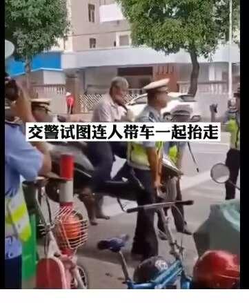 图片记录了街头执法现场：多名身着黄色反光背心、头戴警帽的交警正合力抬抱一名坐在小型车辆（疑似电动车）上的人员，试图将其连人带车一同搬离。周围有手持手机拍摄的围观群众，背景可见树木、居民楼及停放的机动车，画面上方文字标注“交警试图连人带车一起抬走”，直观呈现了执法过程中的肢体互动场景。
