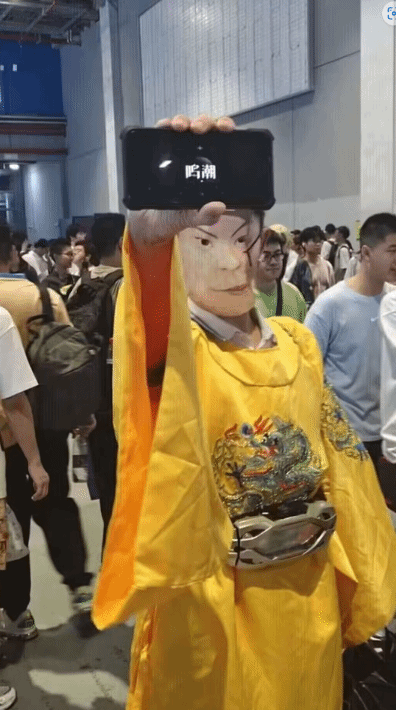这是一张在热闹的线下活动（推测为漫展）现场拍摄的照片，画面中心的人物身着绣有龙纹的黄色传统服饰，模仿古代帝王的着装风格，他将一部智能手机举在自己的脸部正前方，手机屏幕上清晰显示着“鸣潮”的字样，借助视觉错位的巧思，让观看者产生手机就是人物脸部的错觉，制造出诙谐有趣的视觉效果。背景中可以看到大量其他参与活动的人群，大家聚集在一起，烘托出活动现场热闹拥挤的氛围，这是一种结合了游戏元素的趣味整活玩法。
