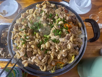 这是一张桑拿鸡的特写照片，展示了一锅热气腾腾的鸡肉料理。鸡肉被切成小块，与葱花和其他配料一起烹饪，盛放在一个带把手的金属锅中。锅的周围可以看到一些餐具和其他食物，整个场景呈现出一种温馨诱人的美食氛围。蒸汽从锅中升起，表明这道菜刚刚做好，非常新鲜。