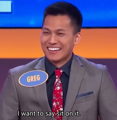 这是一张美国问答综艺《家庭问答》（Family Feud）的截图，图中名为Greg的参赛嘉宾身着灰色西装搭配花红色领带，脸上带着戏谑的大笑表情，画面下方的字幕显示他的发言：“I want to say sit on it.”（我想说‘坐上去’）。这句脱离节目常规答案的无厘头发言，让场景充满滑稽戏谑的效果，成为搞笑梗图在网络流传。