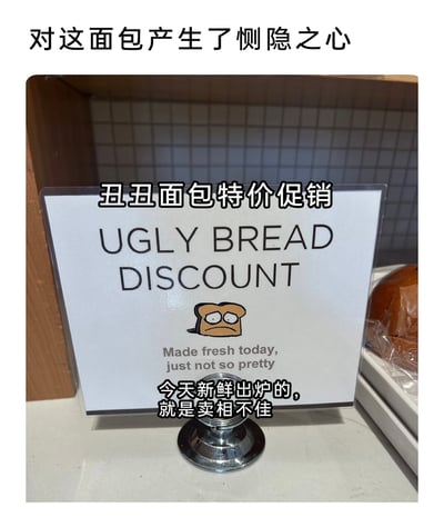 这是一张面包店的实拍图片，画面核心是一个放置在柜台的立牌促销标识。标识上用醒目的英文写着“UGLY BREAD DISCOUNT”，下方搭配了一个画着沮丧、无神表情的卡通面包图案，还有英文小字标注“Made fresh today, just not so pretty”。同时标识上添加了对应的中文翻译：“丑丑面包特价促销”“今天新鲜出炉的，就是卖相不佳”。图片顶部还有用户添加的评论文字“对这面包产生了恻隐之心”，画面右侧能看到包装好的面包。整个内容用幽默又带点共情的方式，推广那些外形不规整但新鲜出炉的面包，既说明了面包的品质没问题，又用趣味文案降低顾客对卖相的顾虑，让人觉得有趣又暖心。