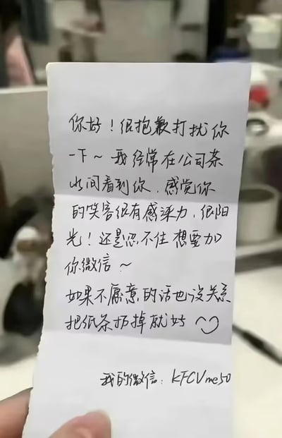 这是一张实拍的手写整活小纸条图片，前半段内容伪装成公司同事故意搭讪，夸赞对方笑容有感染力、性格阳光，提出想要添加对方微信，还贴心表示如果对方不愿意直接扔掉纸条即可，营造出友好交友的预期，最后留下的微信号却是网络流行梗“KFCVme50”，用预期反差制造笑点，属于网友创作的搞笑整活内容。