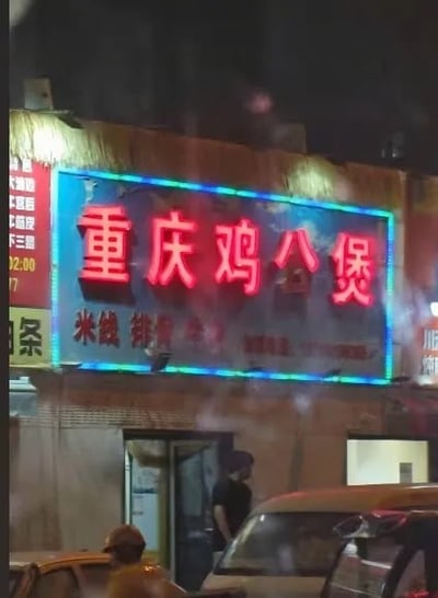 这是一张夜晚拍摄的街头店铺照片，画面中心是一家名为“重庆鸡公煲”的店铺招牌。招牌以红色霓虹灯为主，边缘有蓝色灯光装饰，上面清晰显示“重庆鸡公煲”字样，下方还有“米线”“排骨”等菜品名称。店铺门口有模糊的行人和停放的车辆，整体呈现出城市夜晚街头小吃店的日常景象，灯光温暖，充满生活气息。