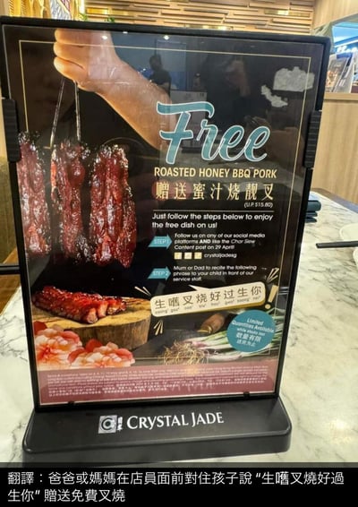 图片展示了餐厅“翡翠拉面小笼包（Crystal Jade）”的促销海报，主打“赠送蜜汁烧靓叉（价值$15.60）”活动。海报左侧是诱人的蜜汁叉烧串特写，右侧为活动说明：顾客需完成两步操作——1. 关注餐厅社交媒体并点赞4月29日的叉烧相关内容；2. 父母需在店员面前对孩子说出“生嚐叉燒好過生你！”（意为“吃叉烧好过生你”）即可获赠。海报底部标注“数量有限，送完即止”。图片下方另有翻译文字：“爸爸或媽媽在店員面前對孩子說‘生嚐叉燒好過生你’贈送免費叉燒”。

**谐音梗/暗示**：中文标语“生嚐叉燒好過生你”利用夸张对比制造话题性，将“生叉烧”与“生孩子”进行幽默（或争议性）比较，通过生活化的调侃吸引顾客注意力。