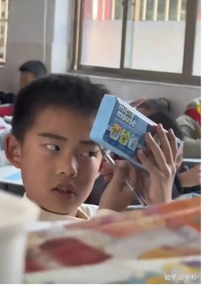 一名学生坐在教室内，举着一个标有"mice mouse"的盒子挡在自己脸旁，似乎在遮挡或做出搞笑动作。这个动作常见于学生之间互相逗趣、应对老师或者伙伴的情境。盒子上的"mice mouse"有点像对“米奇妙妙屋”（Mickey Mouse）的山寨或谐音梗，带来一定幽默效果。整张图片营造出轻松搞笑的校园日常氛围。

【谐音/山寨梗】盒子的品牌疑似山寨知名IP“米奇妙妙屋”。