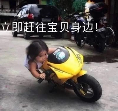 图片中，一名幼儿正俯身骑在一辆黄色的小型玩具摩托车上，双手紧紧握住车把，身体前倾，眼神专注，仿佛即将“火速出发”。背景中可见一辆黑色轿车和一辆摩托车停放在居民楼旁的水泥地面上，环境像是普通的居民区院落。图片上方配有文字“立即赶往宝贝身边!”，通过幼儿认真的小模样与“紧急出发”的文字形成反差萌，营造出既可爱又搞笑的效果，展现了孩子模仿大人骑车的童真场景。