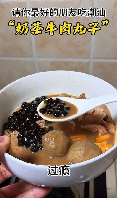 这是一张整活类的美食实拍图，画面中一只手捧着一个白色大碗，碗里原本是传统潮汕牛肉丸汤，包含牛肉丸、肉块、浅褐色的汤底，却被加入了大量奶茶常见的配料黑珍珠，汤勺还舀起了混有珍珠的汤汁，图片顶部配有文字“请你最好的朋友吃潮汕‘奶茶牛肉丸子’”，底部配文“过瘾”，属于将奶茶元素和传统潮汕美食混搭的猎奇创意，带有调侃、整蛊好友的玩梗属性。