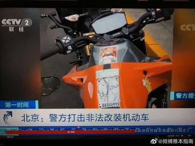 这是央视财经频道（CCTV2）《第一时间》栏目的新闻截图，报道北京警方开展的打击非法改装机动车行动，画面聚焦在一辆橙色摩托车上。该摩托车的车头部位贴有多张低俗风格的二次元动漫贴纸，贴纸内容暴露，违背公序良俗，此摩托车属于此次打击行动中的涉事车辆，新闻意在展示非法改装及违规装饰车辆的整治情况。