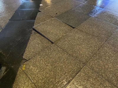 这是一张雨后的户外人行道实拍图，地面的浅棕色花岗岩地砖与深色盲道砖都被雨水打湿，表面带有水渍反光。画面中一块浅棕色地砖明显向上翘起，与周围地砖形成高度差，存在绊倒行人的安全隐患，地面还残留有少量垃圾。