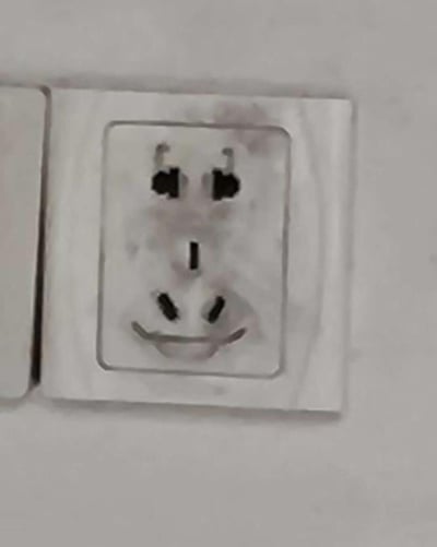 一张日常家庭场景中的插座特写照片，展现了一个有趣的视觉错觉现象。插座的两个上孔自然形成了一双眼睛，中间的接地孔像一个小鼻子，而下边的两个孔则构成了一个微笑的嘴巴，整体看起来仿佛一个带着神秘微笑的脸庞。这种日常生活中的意外发现给普通的家居用品增添了几分趣味性和幽默感。