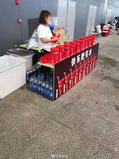 这是活动现场的可口可乐供应摊位，一位工作人员正在将大瓶装可乐分装到红色定制杯中。摊位的宣传板上写着网友对可乐的调侃式爱称“快乐肥宅水”，还配有“红遍全国 爽够一个”“渴乐渴你”的宣传语，下方整齐摆放着多瓶大包装可乐，整体场景接地气又充满轻松的氛围。
其中“快乐肥宅水”是网络流行梗，用来调侃可乐能给人带来简单的快乐，深受网友喜爱。