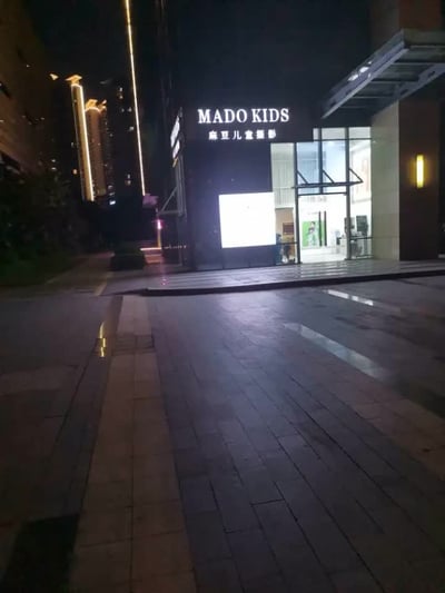 夜晚的商业街区，一栋现代风格建筑的底层商铺亮着灯光，正中央是一家名为"MADO KIDS 麻豆儿童摄影"的儿童摄影店。店铺招牌采用白色发光字体，在深色建筑背景上十分醒目。地面是湿滑的石板路，反射着周围的灯光，营造出雨后或湿润夜晚的氛围。周围有高楼大厦，部分建筑亮着装饰性灯光，整体呈现出城市商业区夜晚的静谧景象。