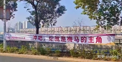这是一张户外实拍的照片，画面主体是一条设置在路边矮墙上的赛事横幅，横幅左侧标注了“广汽埃安·2024梅州马拉松赛”，右侧是醒目的励志标语“冲吧，你就是梅马的主角”。横幅前方有低矮的绿植，背景是错落的城市居民楼和树木，天气晴朗，整体场景传递出对梅州马拉松参赛者的鼓励与支持，没有双关、谐音梗等特殊表意。