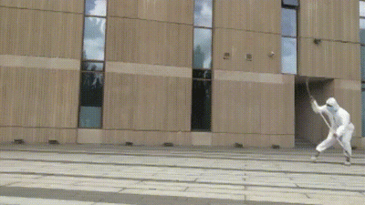这是一段动态GIF，展示了一位身着白色防护服的人在户外建筑前的广场上，手持带刃长柄兵器，依次完成单腿高举支撑、腾空翻转挥舞兵器、稳稳落地的一系列流畅武术动作，防护服的防疫属性与硬核的功夫动作形成有趣反差，展现出十足的力量感与灵活性。