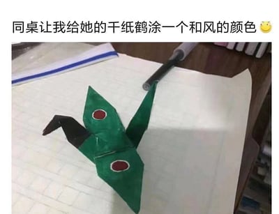 图片展示了一只手工折纸的千纸鹤，主体被涂成绿色，翅膀上画有两个红色圆形图案，看起来像日本军用飞机的样式。图片上方配有文字“同桌让我给她的干纸鹤涂一个和风的颜色😊”。这里存在一个幽默的误解：“和风”本指日本传统风格，但图中的千纸鹤被涂成了类似日本军用飞机（特别是零式战斗机）的配色，形成了有趣的反差萌效果。