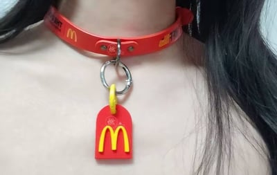 这是一张实拍的创意DIY饰品图，展示了一款由麦当劳周边改造而成的颈圈项圈。项圈主体为红色，印有麦当劳经典黄色M标志和“燃”字，通过金属环悬挂着同款红底黄M的麦当劳挂饰，挂饰上同样印有“燃”字，整体采用麦当劳标志性的红黄配色，将品牌周边与饰品结合，打造出个性潮流的穿搭配饰，无谐音梗或双关含义。