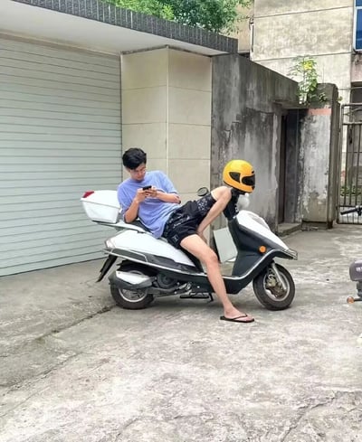 这是一张利用拍摄角度制造视觉错位效果的搞笑真人照片。画面中，一名身穿浅蓝色短袖、黑色花纹短裤、脚踩人字拖的戴眼镜男子，半躺在白色电动车的后座上，低头专注地操作着手机。他的一条右腿弯曲抬起，恰好与电动车车头处的黄色头盔、车把部分重合，从当前拍摄角度看，仿佛电动车“长出”了人的腿，男子像是骑在自己的腿上，形成了极具趣味的视觉错觉。场景位于一处带有灰色车库卷帘门的户外水泥地面区域，背景还有老旧的墙体，整体氛围轻松搞笑，是通过创意拍摄手法打造的趣味日常内容。
