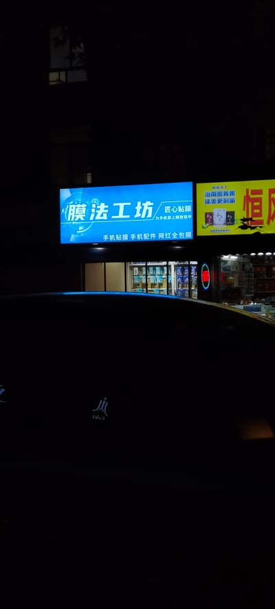 这是一张夜晚户外拍摄的竖版照片，前景是黑色汽车的车顶，背景是亮着灯光的街边商铺招牌。左侧蓝色发光招牌为「膜法工坊」，主打手机贴膜、手机配件与网红全包膜，搭配标语「匠心贴膜，为手机穿上精致铠甲」；右侧黄色招牌是「和成天下」槟榔的广告，标注「海南嫩青果，味美更耐嚼」与「全国高端销量领先」，旁边还有带有「恒」字的店铺招牌露出一角。黑暗的环境让发光招牌显得格外醒目，呈现出日常的街边夜景。