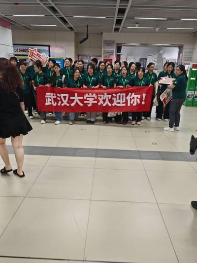 在武汉的地铁站内，一群身着统一深绿色上衣、佩戴绿色挂绳的武汉大学志愿者们集结在一起，共同举着醒目的红色横幅，上面写着「武汉大学欢迎你！」。部分志愿者还手持设计可爱的「你好 新同学」手举牌，整体氛围热情满满，是学校为迎接新入学的新生开展的接站迎新活动，充满了温馨的校园氛围。
无谐音梗、双关或暗示内容。
