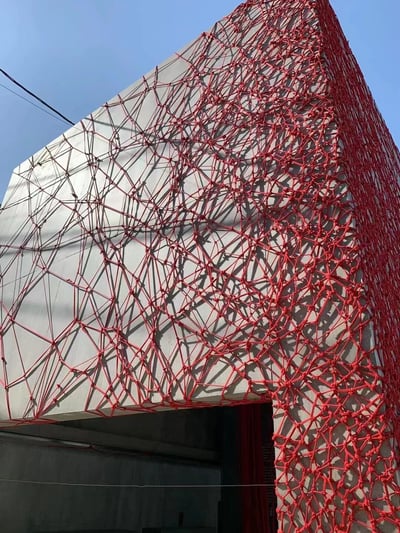 一张展示建筑外观的照片，建筑主体为银灰色，外表面覆盖着密集的红色网状结构，网线交织形成不规则的几何图案。照片从下往上拍摄，背景是晴朗的蓝天，能看到部分电线。