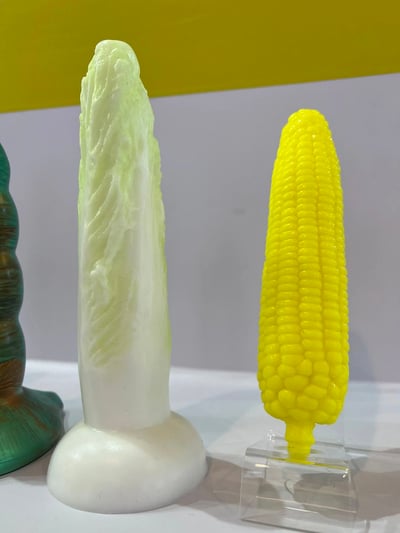 图片展示了两个创意蔬菜造型的摆件。左侧是一个白色的白菜造型雕塑，细节丰富，纹理清晰，底部有一个白色底座。右侧是一个亮黄色的玉米造型摆件，颗粒分明，色彩鲜艳，放置在一个透明的展示架上。背景为黄色和灰色的墙面，整体构图简洁，突出展示了这些有趣的蔬菜造型艺术品。