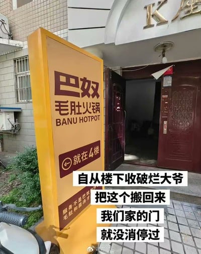这是一张记录现实乌龙事件的搞笑梗图，画面拍摄于居民楼K座的单元门口，门口摆放着一个原本属于巴奴毛肚火锅的黄色立式宣传立牌，立牌上标注有火锅店指引文字“就在4楼”，图片叠加的配文说明该立牌是楼下收破烂的大爷捡回来放在此处的，从此之后住户家经常被误以为这里有火锅店的访客打扰，一直不得安宁，事件充满荒诞的趣味性。