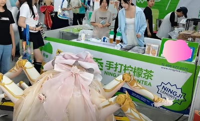 这张图片摄于一次漫展活动现场，一位身穿机甲风格裙装、带有粉色大蝴蝶结的coser蹲或坐在柠季柠檬茶饮品摊前，背景是品牌展台与不少排队买饮品的人群。画面彰显了二次元cosplay文化与商业品牌展会的融汇，具有明显的趣味性和猎奇感，适合分享于动漫圈和线下活动相关社群。