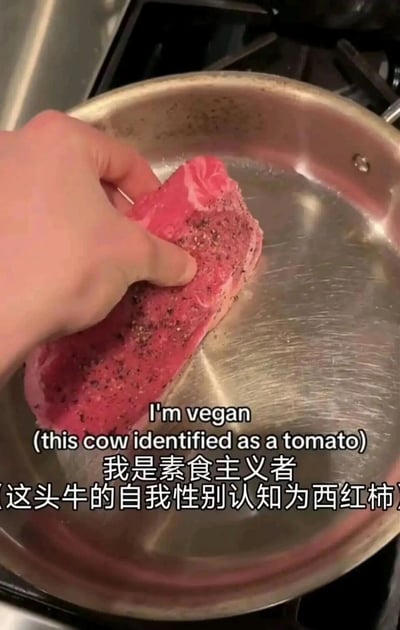 这是一张带有搞笑加注文字的美食整活静态图，画面中一只手将一块撒了黑胡椒和食用盐、外观酷似生牛排的红色西红柿放置在不锈钢平底锅上，搭配的中英文文字玩了一个荒诞的身份认知梗：英文标注“I'm vegan (this cow identified as a tomato)”，中文翻译为“我是素食主义者（这头牛的自我性别认知为西红柿）”，通过把伪装成牛排的西红柿调侃成“自我认知为西红柿的牛”，以幽默的方式化解素食主义者无法食用肉类的限制，制造出反差感拉满的搞笑效果，属于趣味美食创作类的玩梗内容。